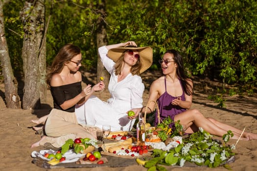Three cheerful girlfriends at a summer picnic. Happy women drink wine in nature. Vacation of free girls of Caucasian appearance in the summer on the sand