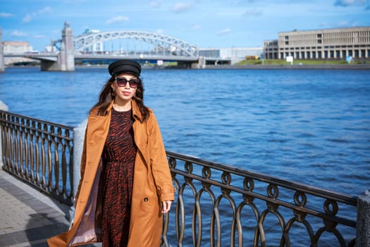 A young European woman in sunglasses and a black cap poses on the embankment in a brown coat on a spring sunny day. The concept of a successful and free woman