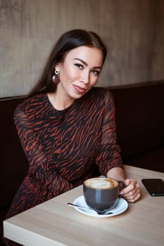 A young beautiful woman with dark hair, Caucasian appearance with makeup in a brown dress is sitting at a table in a cafe with coffee and smiling with a snow-white toothy smile