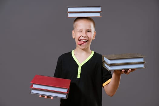 Funny Caucasian boy holding books in his hands and on his head. Shows her tongue on a gray background in casual wear. Back to school concept