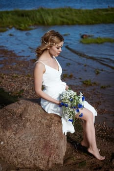 A young woman alone, in a white dress, sits on a stone on the shore, holding a bouquet in her hand. In summer in sunny weather
