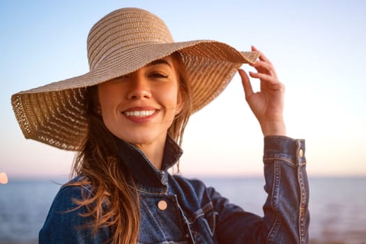 Portrait of a happy elegant young woman in a white denim jacket and a straw hat on the ocean shore at sunset, looking into the distance. Young caucasian female model close up on the seashore.