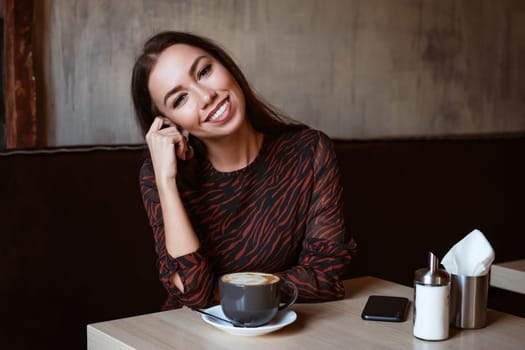 Portrait of a young woman of Caucasian appearance in a cafe with a cup of coffee. Beautiful brunette with brown eyes. Enjoying morning coffee in a cozy cafeteria