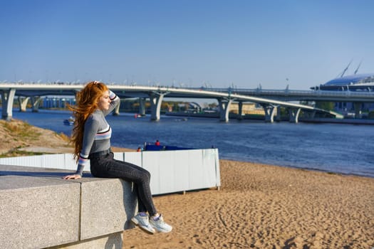 Beautiful young woman with long hair of Caucasian nationality , in casual clothes sits on the embankment of the river on a spring sunny day with a view of the bridge