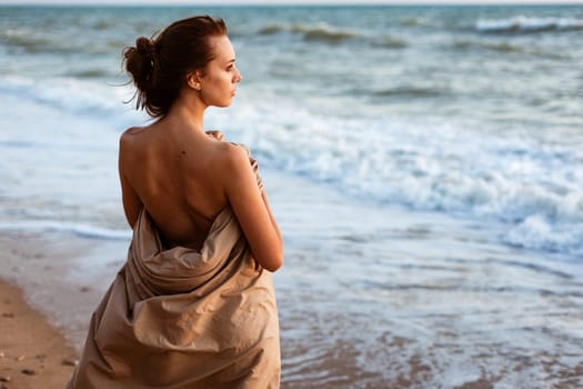 The girl on the seashore in the evening stands. Lonely looking into the distance on the beach at sunset. Summer evening by the water. Beautiful young woman wrapped in a blanket.