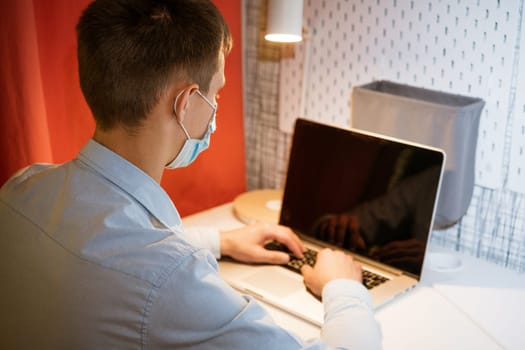 a young Caucasian man in a light shirt sits at a table in the evening at home wearing a protective medical mask and a mask and works at a laptop, remote work at home online