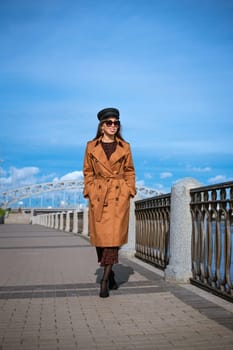 Beautiful caucasian woman in sunglasses, in a black cap and jacket posing while standing on the embankment on a sunny day against a background of blue sky and cityscape