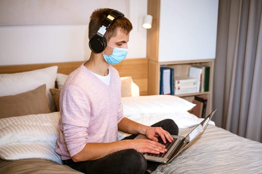 a young man of european appearance in casual clothes sits on a bed in a room at home wearing headphones and a protective mask at home with a laptop, the concept of remote work and education