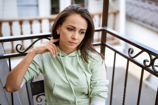 pensive young brunette woman in casual clothes sits in a chair on the balcony in summer