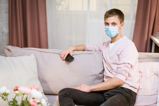 young caucasian man sitting on a sofa in casual clothes, wearing a protective medical mask with a phone in his hand