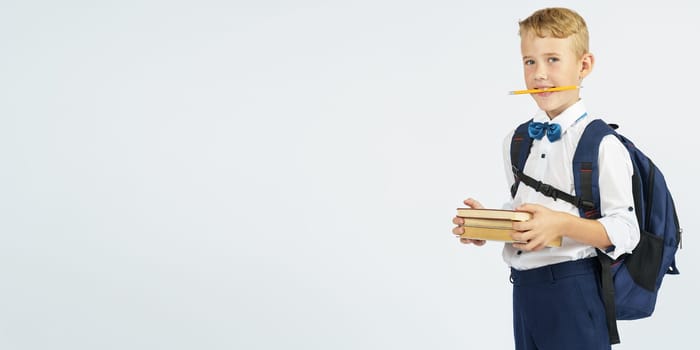 A schoolboy with a briefcase holds books and a pencil in his teeth. Looks at them. Isolated background. Education concept