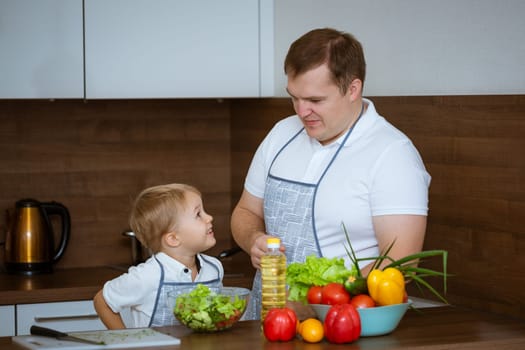 The blonde-haired son and father prepare a salad of colorful, fresh vegetables together in the kitchen. They both wore blue aprons.