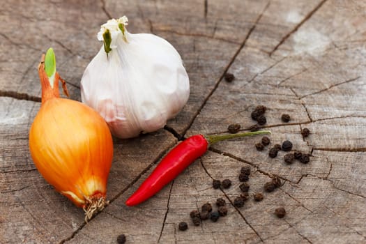 a head of garlic, onion and red hot pepper on a cracked wooden stump background close-up on a sunny summer day. the row is sprinkled with black pepper peas