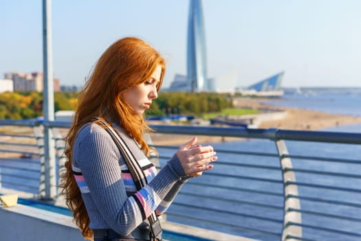 Young red-haired woman of Caucasian ethnicity , with a phone in her hand stands on a bridge overlooking the city on a sunny spring day looks into the phone