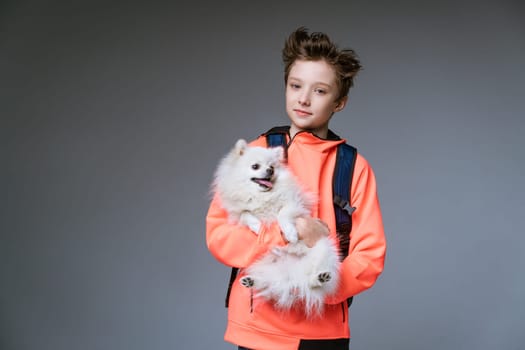 cheerful cute schoolboy of Caucasian ethnicity in bright clothes with a backpack and a dog in his hands on a gray background posing.