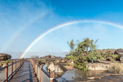 A rainbow is visible over a boardwalk at the Augrabies waterfalls in the Orange River. The river is in flood