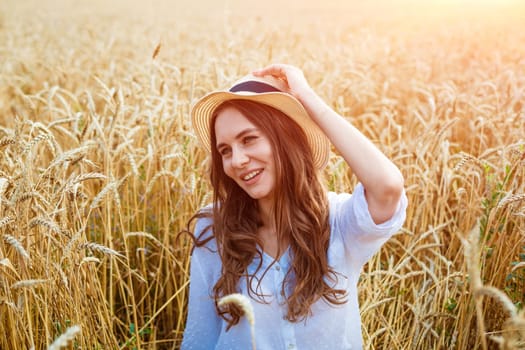 Happy girl sits in wheat field in straw hat. Cute young woman of Caucasian ethnicity in casual clothes enjoys ripe golden wheat in a field. Free woman concept