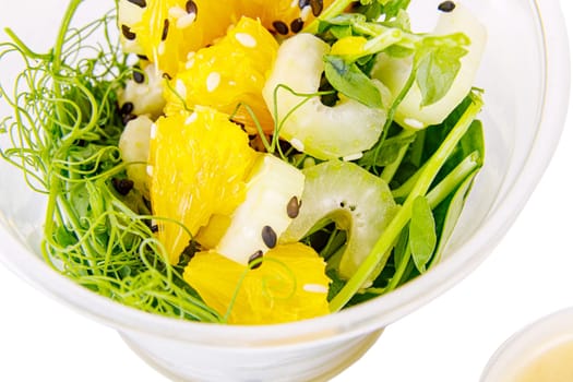 Salad with microgreens peas, spinach, celery, orange, sesame seeds and dressing in plastic cup on white background