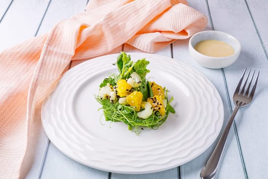Salad with microgreens of peas, spinach, celery, orange, sesame seeds and dressing in plate on white wooden background