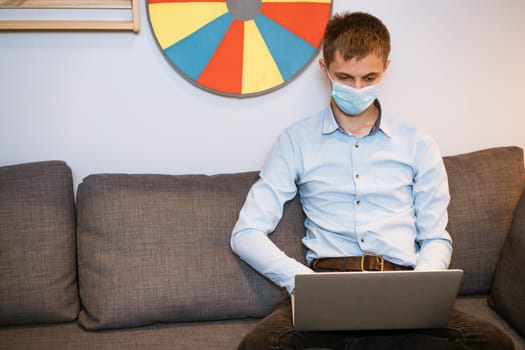 a young guy of European appearance in a light shirt, sits in a protective medical mask and works on the couch via video communication, remote work during a global pandemic