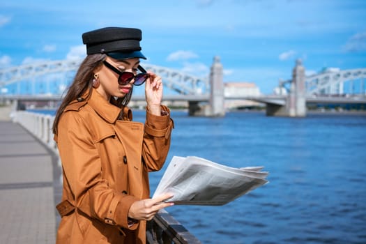 Stylish young woman of Caucasian ethnicity reads fresh newspaper in sunglasses and black cap. Beautiful brown coat standing on embankment of river on sunny day against background of sky and bridge