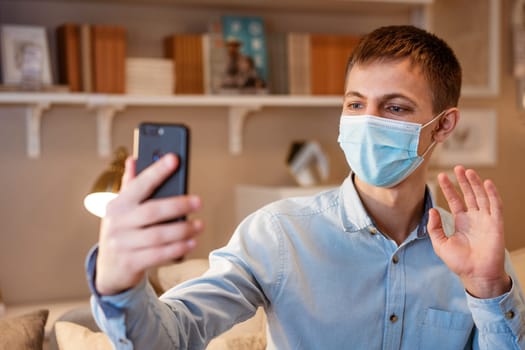 young man of European appearance in casual clothes, at home on the couch in a medical mask communicates by video communication on the phone, the concept of remote online communication on the Internet