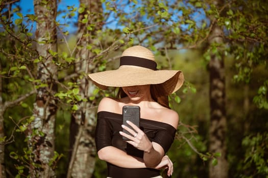 Beautiful happy young woman of caucasian ethnicity in a straw hat from the sun with large brim in a black dress with a phone in her hand smiling on a sunny summer day