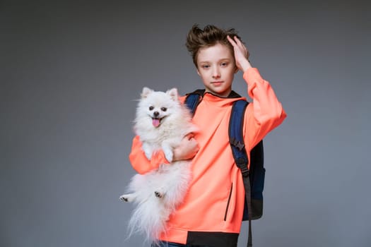 cheerful cute schoolboy of Caucasian ethnicity in bright clothes with a backpack and a dog in his hands on a gray background posing.