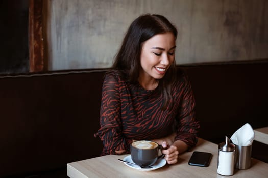 Cheerful girl of Caucasian ethnicity sits at a table in a cafe with a cup of coffee. Beautiful young brunette woman with a beautiful make-up and a white-toed smile smiling