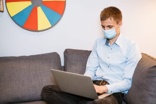 a young man of Caucasian appearance in a blue shirt and a protective mask with a laptop on the couch works at home. Remote work in quarantine