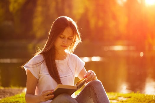 Young caucasian woman sitting on the shore of the lake reads a book on a sunny day. Reads outdoors in the park