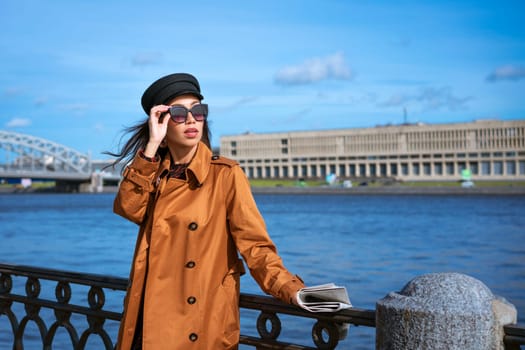 A female portrait of European appearance on the embankment on a sunny spring day with a newspaper in her hand. Beautiful brunette woman in sunglasses and in a brown jacket and black cap