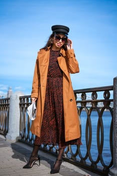 A female portrait of European appearance on the embankment on a sunny spring day with a newspaper in her hand. Beautiful brunette woman in sunglasses and in a brown jacket and black cap