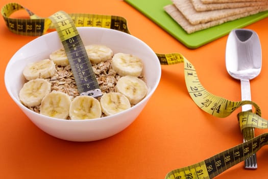 oatmeal with sliced banana and crispbread lie with spoon and yellow measuring tape on orange background, healthy food and diet concept