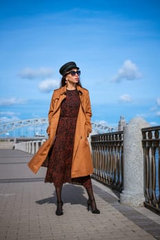 Beautiful young woman of caucasian appearance in a brown raincoat and dress posing on the embankment on a sunny day