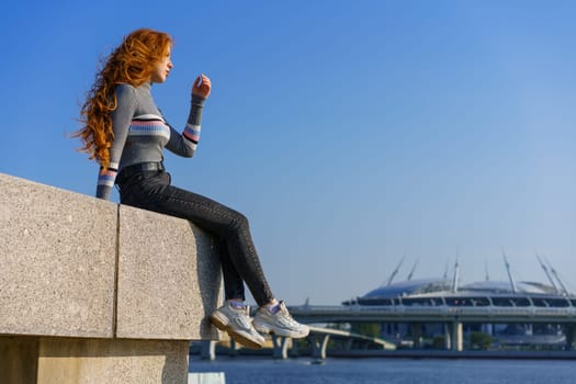 Beautiful young woman with long hair of Caucasian nationality , in casual clothes sits on the embankment of the river on a spring sunny day with a view of the bridge