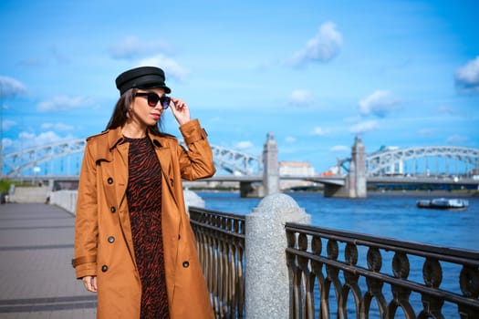 Happy young woman of Caucasian ethnicity in a light brown coat and black cap posing on the embankment against the backdrop of the bridge and the blue sky.