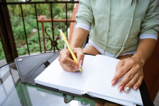 woman writes in a notebook with a pen while sitting at the table at home on the terrace