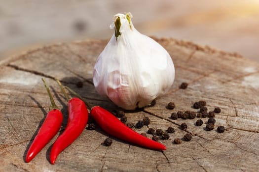 a head of garlic and red hot pepper on a cracked wooden stump background close-up on a sunny summer day. the row is sprinkled with black pepper peas