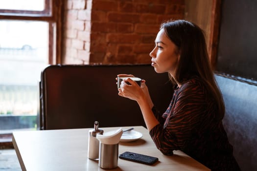 A young beautiful woman of Caucasian appearance sits at a table with a mug of coffee by the window in a cafe, looking to the side with a pensive look