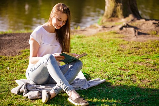 Young caucasian woman sitting on the shore of the lake reads a book on a sunny day. Reads outdoors in the park