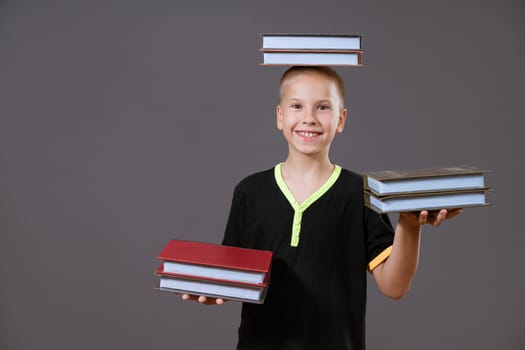 Funny Caucasian boy holding books in his hands and on his head. Shows her tongue on a gray background in casual wear. Back to school concept