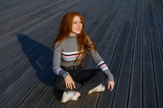 Happy young woman with long red hair of Caucasian nationality, is sitting in casual clothes in the park on a wooden deck on a sunny day and smiling