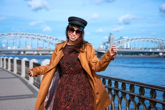 Happy young woman of Caucasian ethnicity in a light brown coat and black cap posing on the embankment against the backdrop of the bridge and the blue sky.