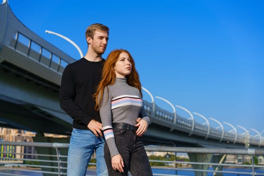 happy young couple man and woman with long red hair, stand against the background of a blue sky and a bridge in casual clothes and smile. Cheerful guy and Caucasian girl on a sunny day