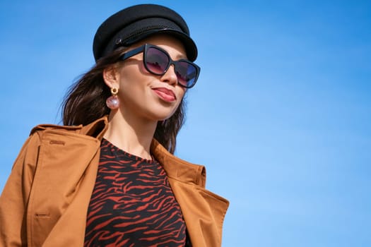 Female portrait of a beautiful young woman of Caucasian appearance in a black cap against a blue sky in sunglasses and a jacket