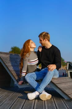 happy couple a guy and a girl with long red hair are sitting on a wooden deck in an embrace. Young man and woman of Caucasian ethnicity in casual clothes on a warm sunny day hugging