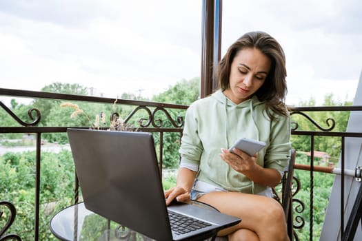 A young woman in home clothes sits on the balcony, at a table works with a phone at a laptop in the summer afternoon