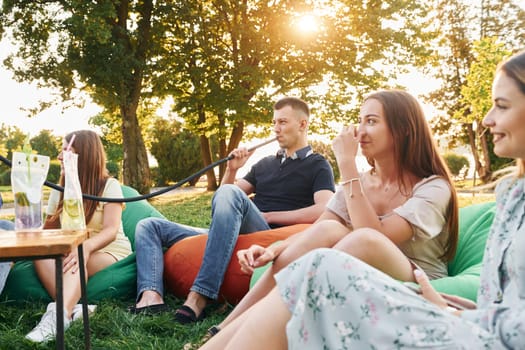 Sitting in the armchair bags and smoking hookah. Group of young people have a party in the park at summer daytime.
