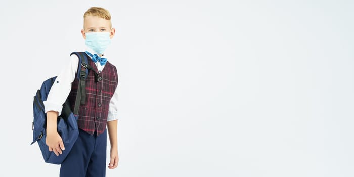 A schoolboy with a medical mask on his face holds a school backpack, stands sideways, looks at the camera. Education concept